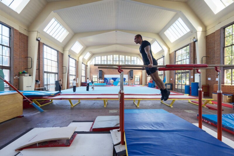 A gymnast performs a skill on parallel bars inside a spacious gymnasium with high ceilings and large windows. Other athletes and gymnastics equipment are visible in the background.