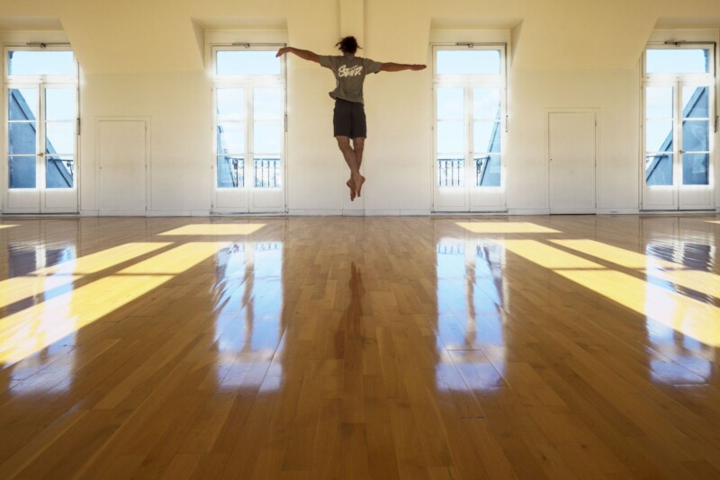 A person jumps with arms outstretched in the center of a sunlit dance studio with large windows and polished wooden floors. Shadows create patterns on the floor, enhancing the sense of movement and space.