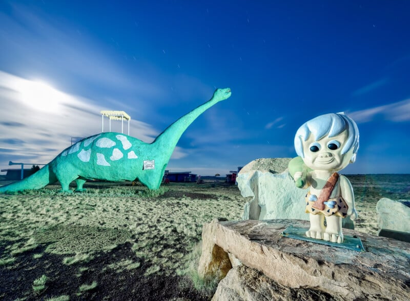 A turquoise dinosaur sculpture and a cartoon-like stone age child figure stand in a grassy outdoor area under a partly cloudy sky. The dinosaur has a long neck, and the child holds a club, wearing a spotted outfit.