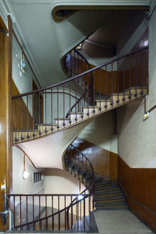 A complex, multi-story spiral staircase inside a building, featuring wooden railings and paneled walls. The stairway twists and turns, leading up through several levels, with lighting casting shadows on the walls.