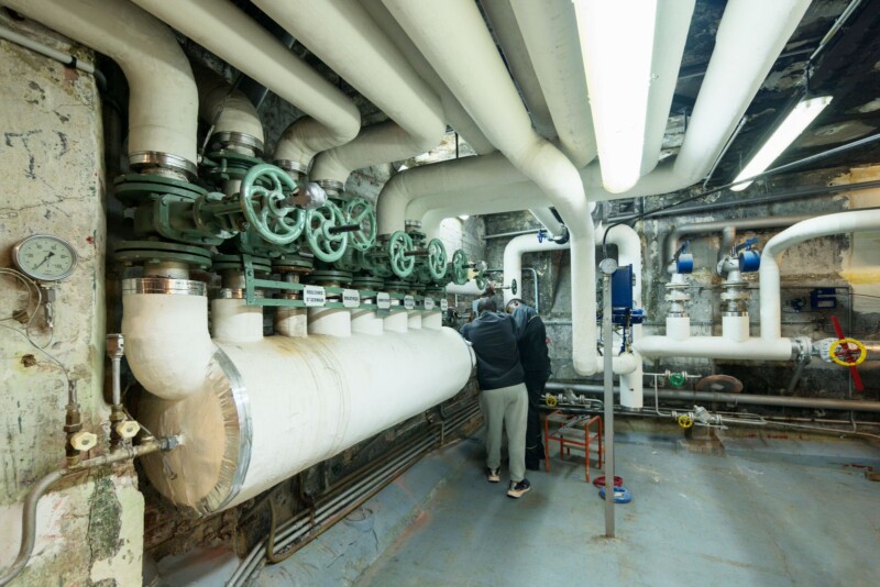 A person working on a network of large, white insulated pipes with green valves in an industrial setting. The room is lit by fluorescent lights, and the floor is concrete. The walls are worn and a small stool is nearby.