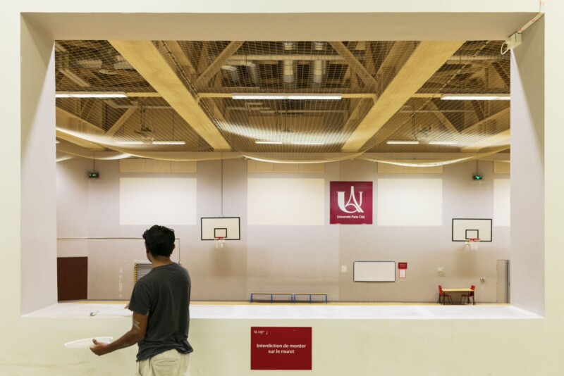 A person stands facing a gymnasium, viewed through a large indoor window. The gym has basketball hoops and a logo for a university. The ceiling features exposed beams and netting. The person holds a plate and wears a dark shirt.