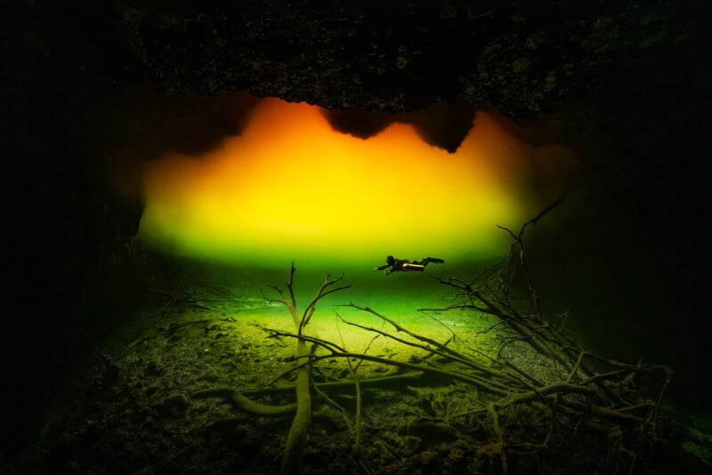 A diver swims through a mysterious underwater cave, illuminated by a gradient glow of yellow and green light. The cave floor is scattered with branches, and the rocky ceiling adds to the enigmatic atmosphere.