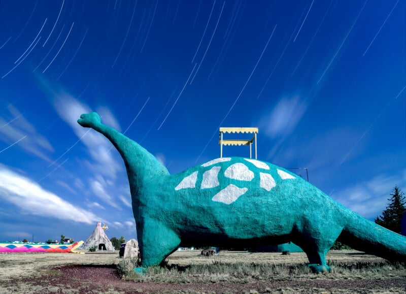 A large green dinosaur sculpture with white markings stands under a starry night sky with star trails. A teepee and a small building are visible in the background. The scene is illuminated with long-exposure lighting effects.