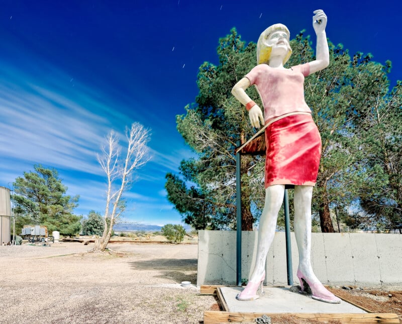 A tall, colorful statue of a woman wearing a pink top and red skirt stands in a desert landscape. She is raising her right hand, set against a backdrop of trees, a cloudy sky, and a barren tree to the left.