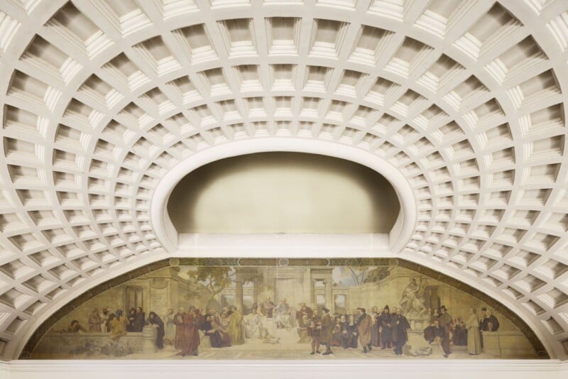 Ornate coffered ceiling with a large, arched mural depicting historical figures in a classical setting. The painting includes a mix of people, trees, and architectural elements, framed by the intricate design of the ceiling.