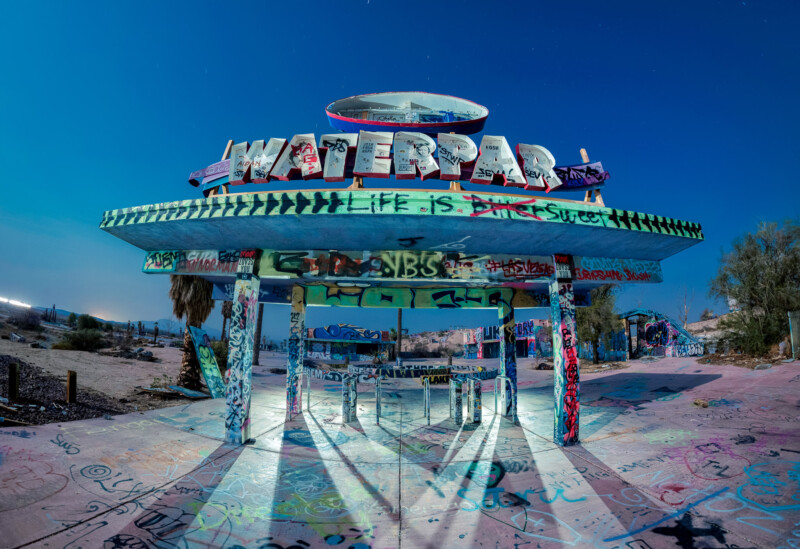 Deserted water park entrance covered in colorful graffiti with a large sign reading "WATERPARK." The structure is set against a clear blue sky, surrounded by arid landscape. Shadows stretch across the ground.