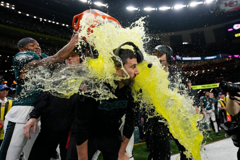 A football coach on the field is being splashed with a cooler of yellow sports drink by players from his team. He is wearing a headset, and the players are celebrating with enthusiasm. Football players and equipment are visible in the background.