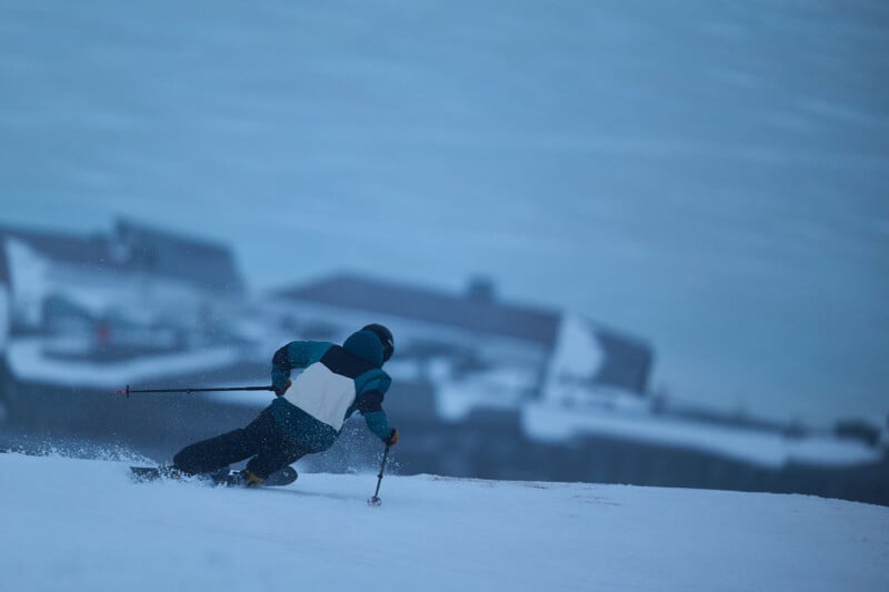 A skier in a blue and white jacket and helmet navigates down a snowy slope, leaving a trail of snow in the air. The background is blurred, showing indistinct buildings or structures against a wintry sky.