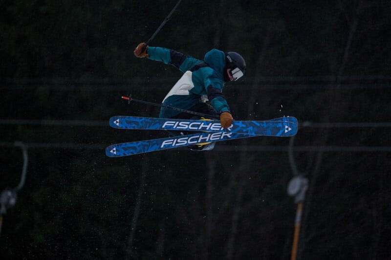 A skier in mid-air performs a trick with blue Fischer skis, wearing a blue jacket, black helmet, and goggles. The background is dark and blurred, suggesting motion and speed.