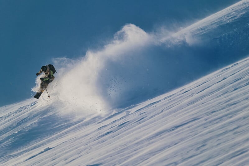 A skier in mid-air navigates a snowy slope, kicking up a wave of powder against a clear blue sky. The dynamic motion captures the thrill and freedom of skiing in a winter landscape.