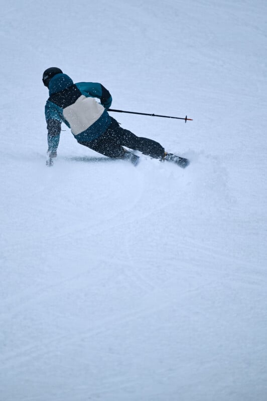 A skier in a blue and white jacket and black pants makes a sharp turn on a snowy slope, with snow spraying up from the skis. The skier is leaning to one side and holding ski poles, creating an action-packed scene.