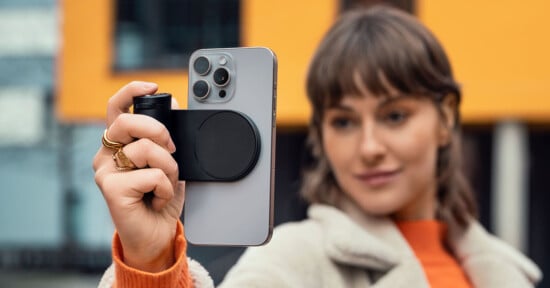 A person holds a smartphone with a camera accessory while taking a selfie. They are wearing a beige coat and an orange top. The background features a blurred yellow and black building.