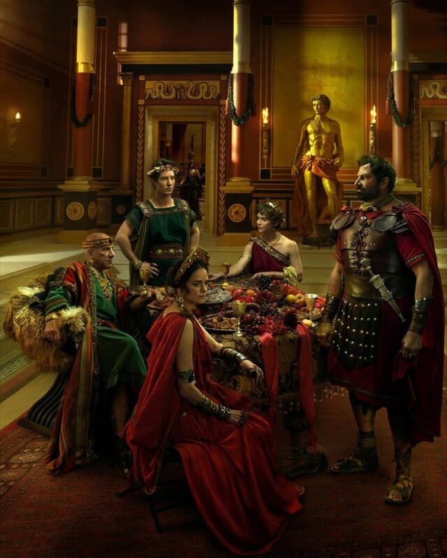 A group of people in ancient Roman attire gather around a table laden with fruit. They are in a grand room with pillars, elaborate decorations, and a large golden statue. The setting conveys a sense of luxury and history.