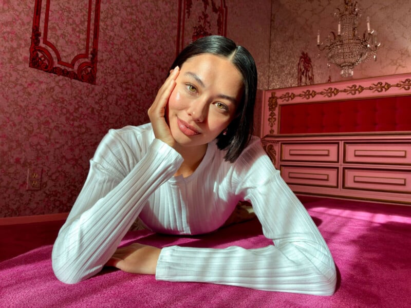 Person with short hair, wearing a white ribbed top, is lying on a pink carpet. They are resting their head on one hand and smiling at the camera. The room has ornate pink wallpaper and a chandelier.