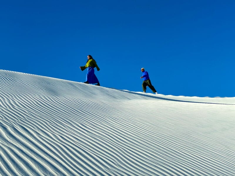 Two people walk along the crest of a white sand dune under a clear blue sky. The surface of the dune has gentle ridges, adding texture to the scene. One person wears a green shawl, and the other is in a blue jacket.