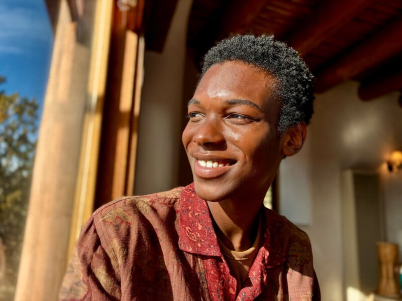 A person with short curly hair smiles while sitting indoors, bathed in warm sunlight. They are wearing a patterned shirt, and the background includes a large window with natural light streaming in.