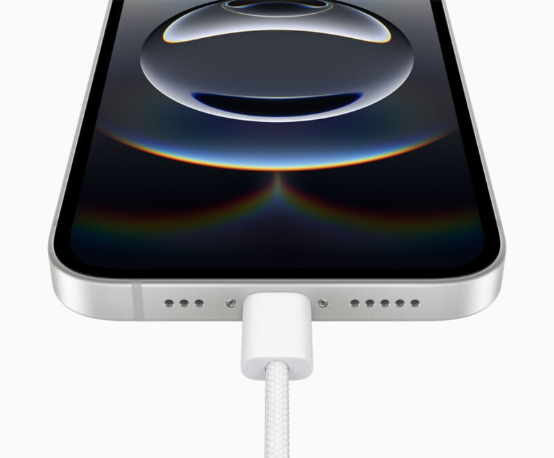 Close-up of a smartphone being charged with a white USB-C cable. The phone screen is on, displaying a spherical pattern with dark and light gradients. The edge of the phone has a sleek, metallic finish.