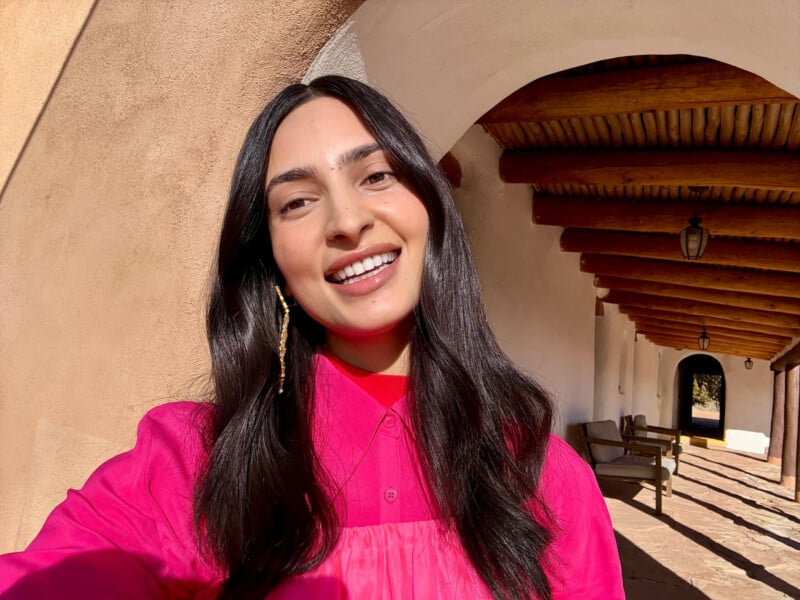 A person with long dark hair smiles in a selfie. They wear a bright pink top and a dangling earring. The background features an arched hallway with wooden beams and a hanging lantern.