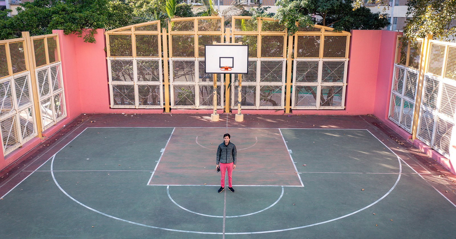 A person stands on a basketball court, centered under the hoop. The court is surrounded by tall fences with plants visible beyond them. The surface is marked with white lines and shows some leaf debris. The person wears a black jacket and red pants.