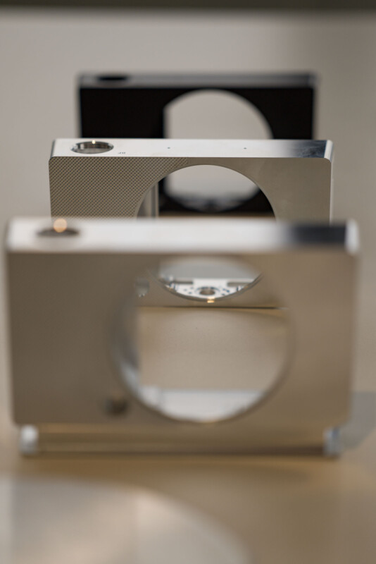 Close-up of three metal camera bodies with circular cutouts, aligned in a row. The bodies have a sleek, minimalist design, with the front one in focus and the others blurred, highlighting the texture and curves of the metal surfaces.