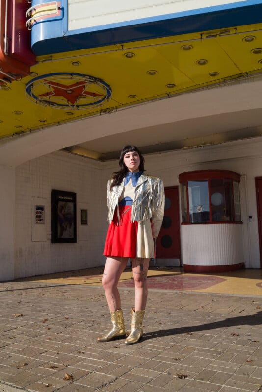 A person stands in front of a retro theater entrance, wearing a metallic fringed jacket, red skirt, and gold boots. Yellow marquee signage is above, and the ground is paved with patterned bricks. The weather appears sunny.