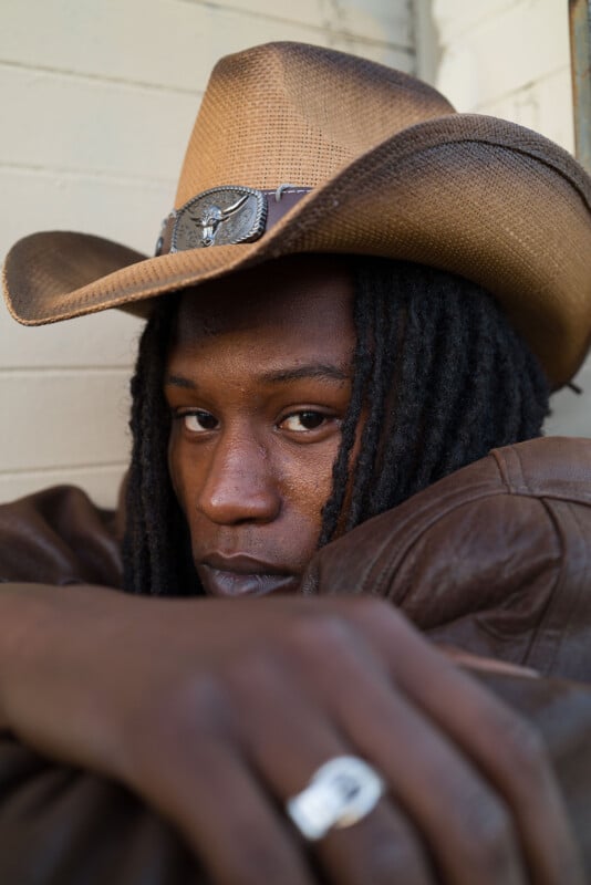 A person with long black hair and a cowboy hat gazes intently at the camera. They are wearing a brown leather jacket and a ring with a large stone, resting their arms in front. The background is a white painted brick wall.