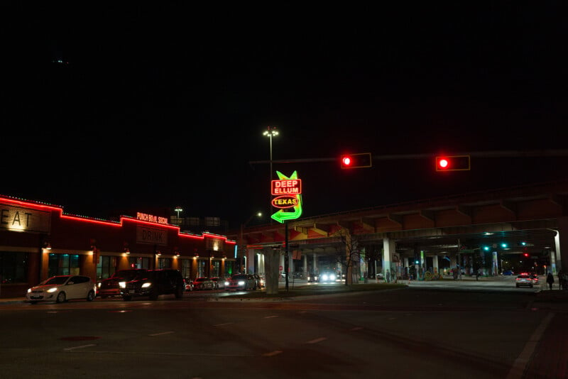 Nighttime street scene in Deep Ellum, Texas, featuring illuminated red and green neon signs. Buildings on the left display signs with words like "DRINK." A bridge spans the road with cars stopped at a traffic light. Streetlights illuminate the area.