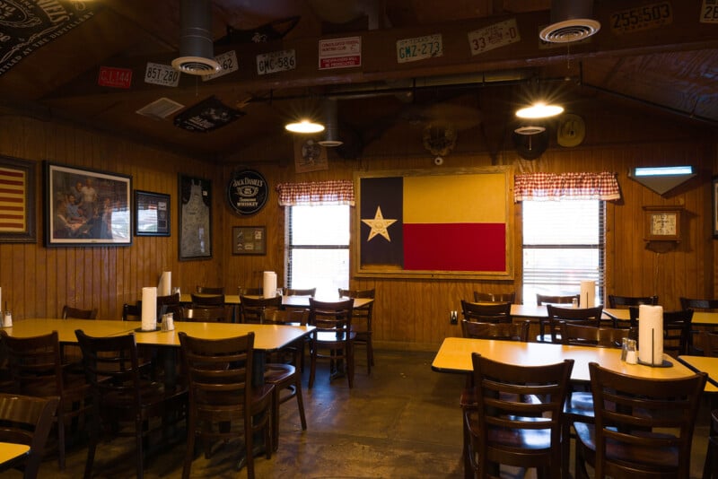 The image shows an empty restaurant with wooden walls and tables. A large Texas flag hangs on the wall, surrounded by various decorative items like license plates and framed pictures. The lighting is warm and inviting.