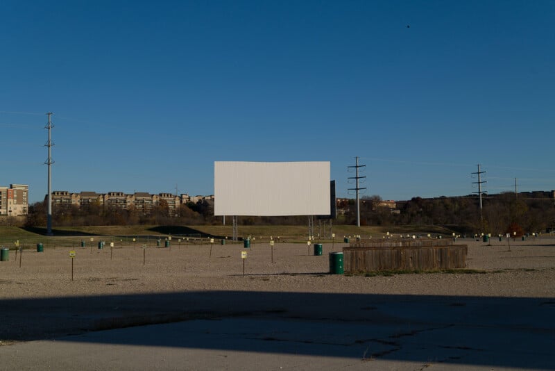 A large empty drive-in theater with a blank screen and open gravel lot. Utility poles are scattered around. Buildings and tree-lined hills are visible in the background under a clear blue sky.