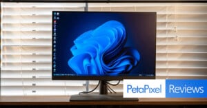 A desktop computer monitor displaying a swirling blue abstract design on its screen. The monitor is placed on a wooden table in front of white blinds. The image has the text "PetaPixel Reviews" overlaid in the bottom right corner.