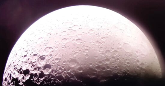 Close-up image of the moon’s surface capturing the cratered terrain. The illuminated portion reveals various sizes of craters and rough textures. The top of the moon is brightly lit, gradually darkening toward the bottom edge.