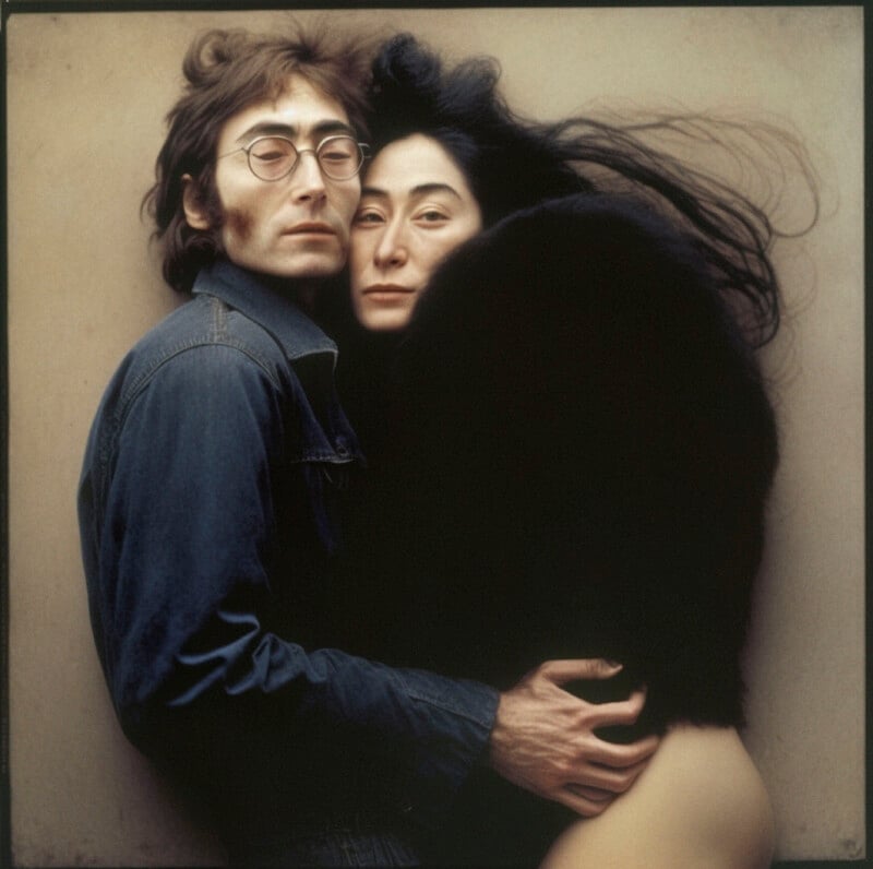 A man wearing glasses and a denim jacket embraces a woman draped in a black garment. Her long black hair flows outward. They both gaze solemnly at the camera against a neutral background.