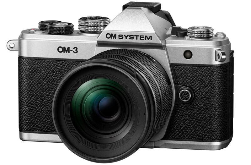 A silver and black camera with a textured grip and multiple knobs on top. The camera has a large lens and the brand name "OM SYSTEM" displayed near the top.