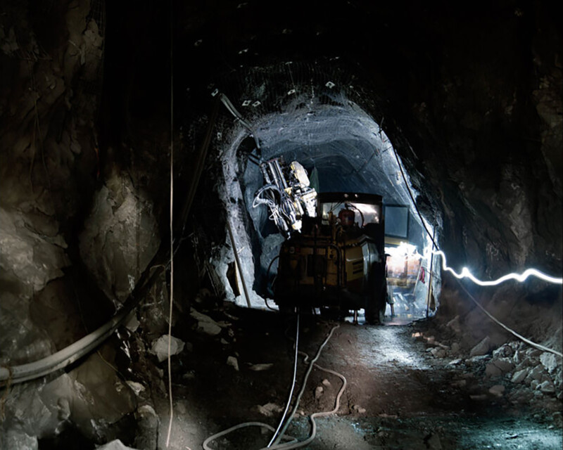 A dimly lit underground mine tunnel with a mining vehicle in the center. The vehicle is equipped with drilling machinery, and there are cables and pipes along the tunnel walls. The environment appears rocky and industrial.