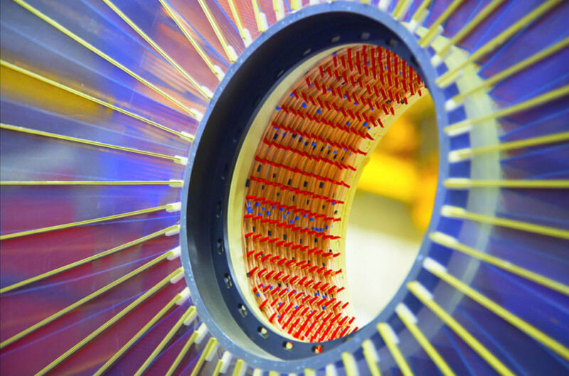 Close-up of a cylindrical structure with red and blue components. The central circular opening is framed by yellow and blue linear elements, creating a dynamic, radial pattern extending to the edges of the image.