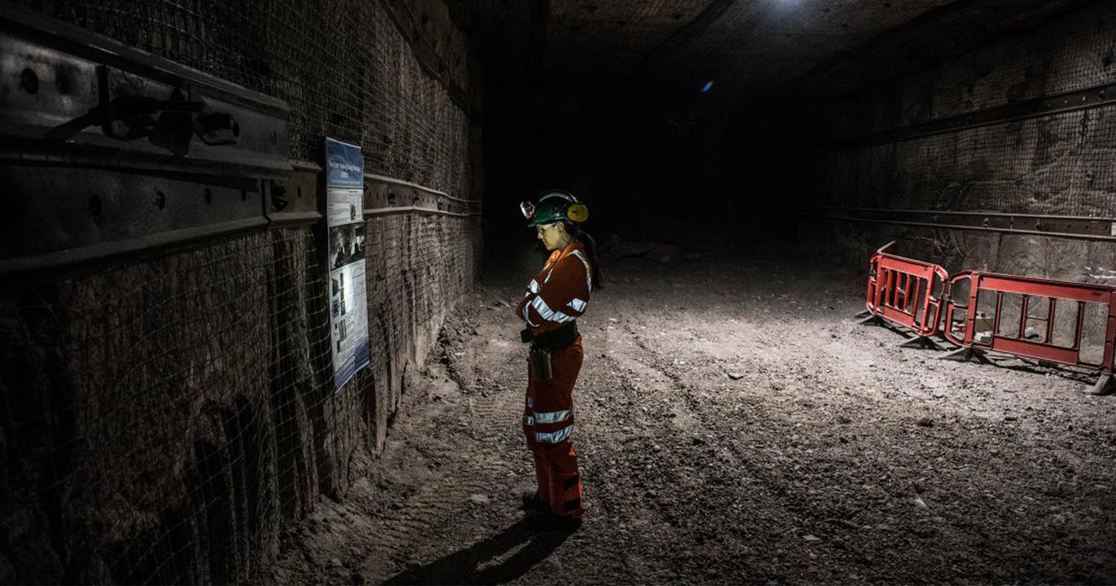 A person in a red protective suit and helmet stands in a dimly lit underground tunnel, reading a sign. The tunnel walls are reinforced, and safety barriers are visible in the background.