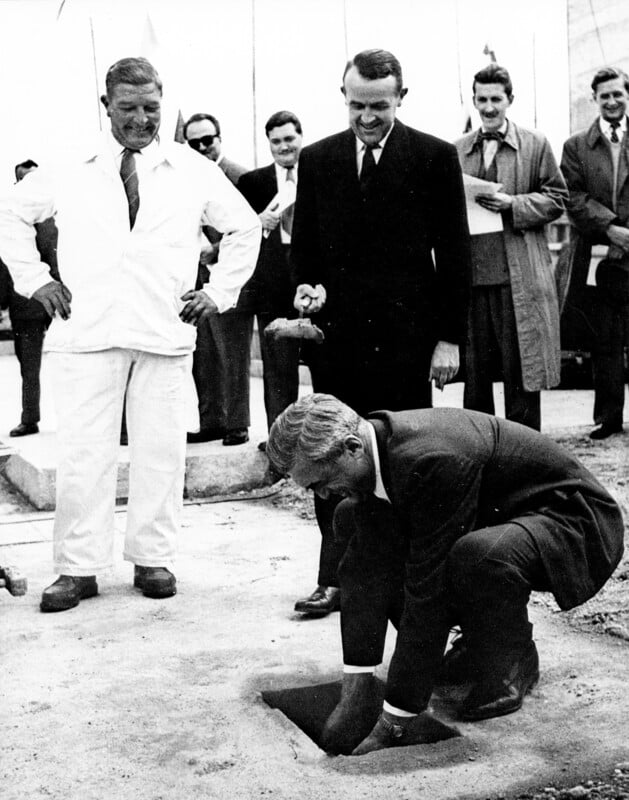 A man in a suit kneels, placing an object into a square hole in the ground, watched by a smiling crowd of men in suits and coats. Another man stands nearby, also holding an object. The setting appears ceremonial.