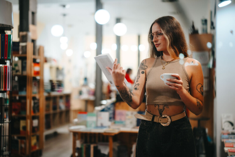 A woman with tattoos and glasses stands in a bookstore, wearing a sleeveless top and holding a white mug. She is looking at a book in her hands. Shelves filled with more books and soft lighting are in the background.
