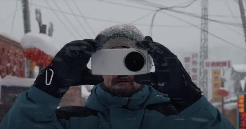 A person in winter clothing and gloves stands in a snowy setting, holding a camera or phone up to take a picture. Snow covers their hat, and there are buildings and power lines visible in the background.