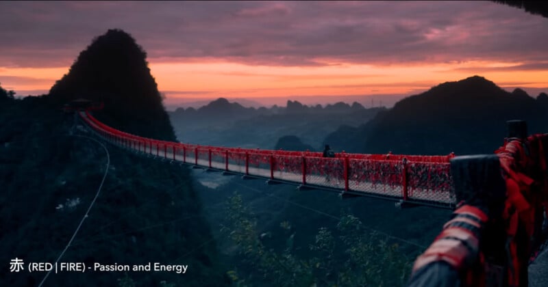 A long red suspension bridge stretches across a mountainous landscape at sunset. The sky is painted in shades of pink and orange, with silhouetted peaks in the background. The bridge is lined with red fabric. Text reads: "赤 (RED) [FIRE] - Passion and Energy.