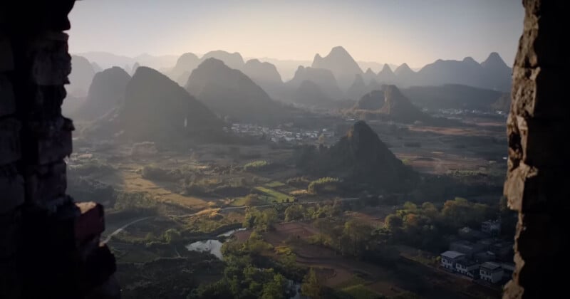 A panoramic view from a window reveals a valley with lush greenery and scattered buildings. In the background, rugged karst mountains rise under a clear, hazy sky, suggesting an early morning or late afternoon setting.