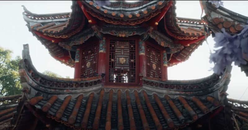 A traditional East Asian pavilion with ornate, multi-tiered curved roofs and intricate wooden carvings. The structure is set against a clear sky, and some foliage is visible around it.