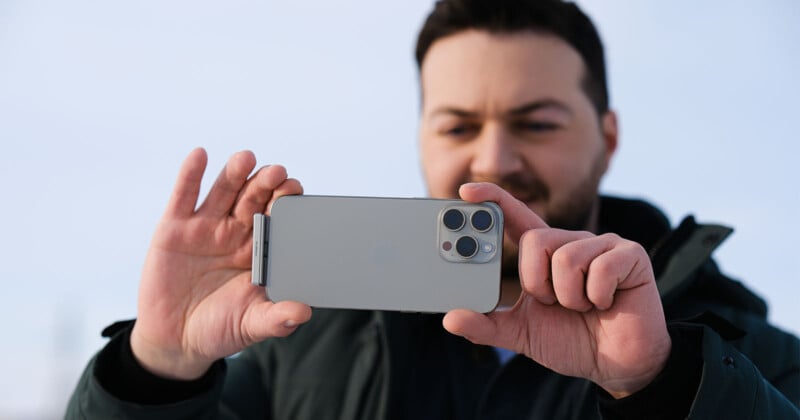 A person is holding a smartphone horizontally, using it to take a photo or video. The person is wearing a dark jacket and standing in an outdoor setting with a clear sky visible in the background.