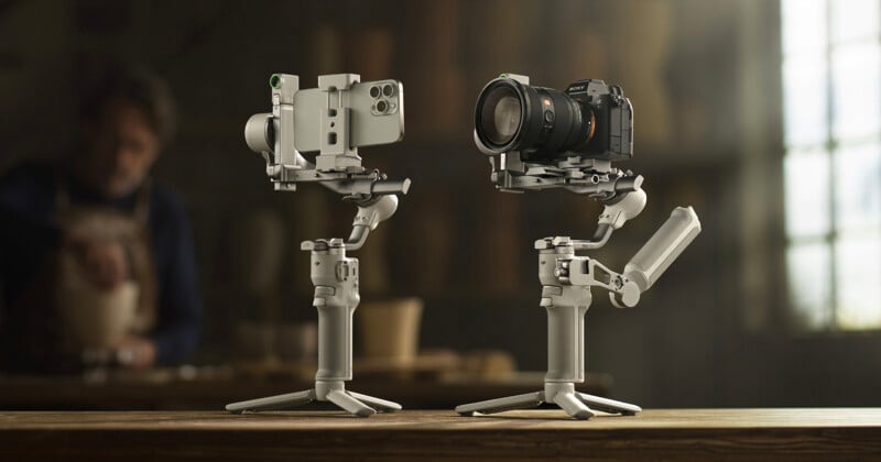 Two gimbals with mounted cameras are displayed on a wooden table. One gimbal holds a smartphone, the other a DSLR camera. A blurred person is in the background, creating a focused scene on the equipment.