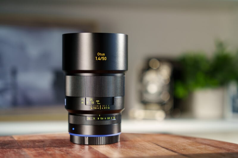 A sleek, black camera lens labeled "Otus 1.4/50" sits on a wooden surface. The background is softly blurred, revealing hints of green from a plant and a framed picture, emphasizing the lens as the focal point.