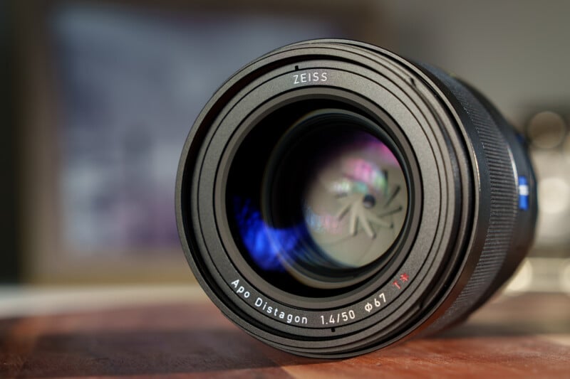 Close-up of a Zeiss camera lens, displaying the text "Apo Distagon 1.4/50 Ø67 T*" around the outer edge. The lens reflects blue light highlights, suggesting depth and precision. The background is softly blurred.