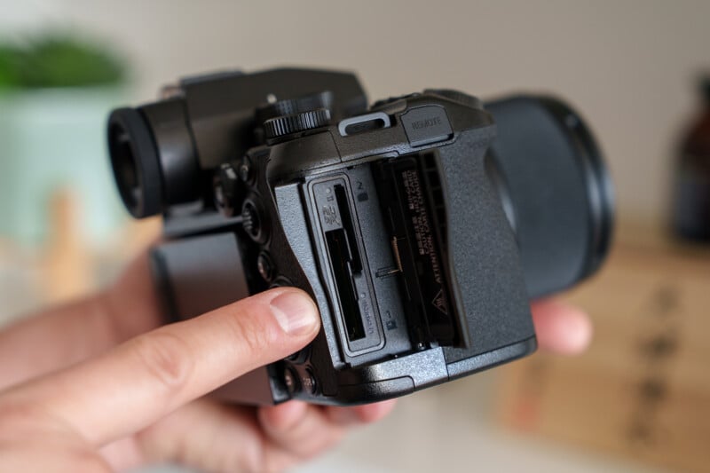 A person holding a black DSLR camera with the SD card slot cover open, showing the SD card slot. The person's fingers are touching the camera, and the background is softly blurred.