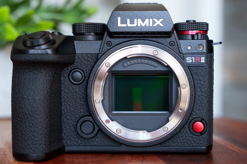 Close-up of a black Lumix S1R II camera body without a lens, showing the sensor. It features textured grip, multiple dials, and buttons. The background is blurred, with hints of a plant visible.