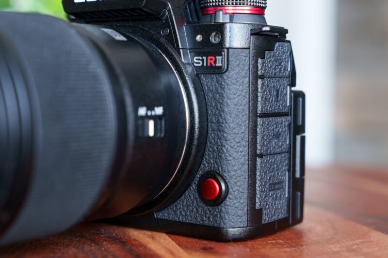 Close-up of a black camera with the model "S1R II" visible near the lens. A red button is prominent on the textured surface of the camera body. The background is blurred wood and greenery.